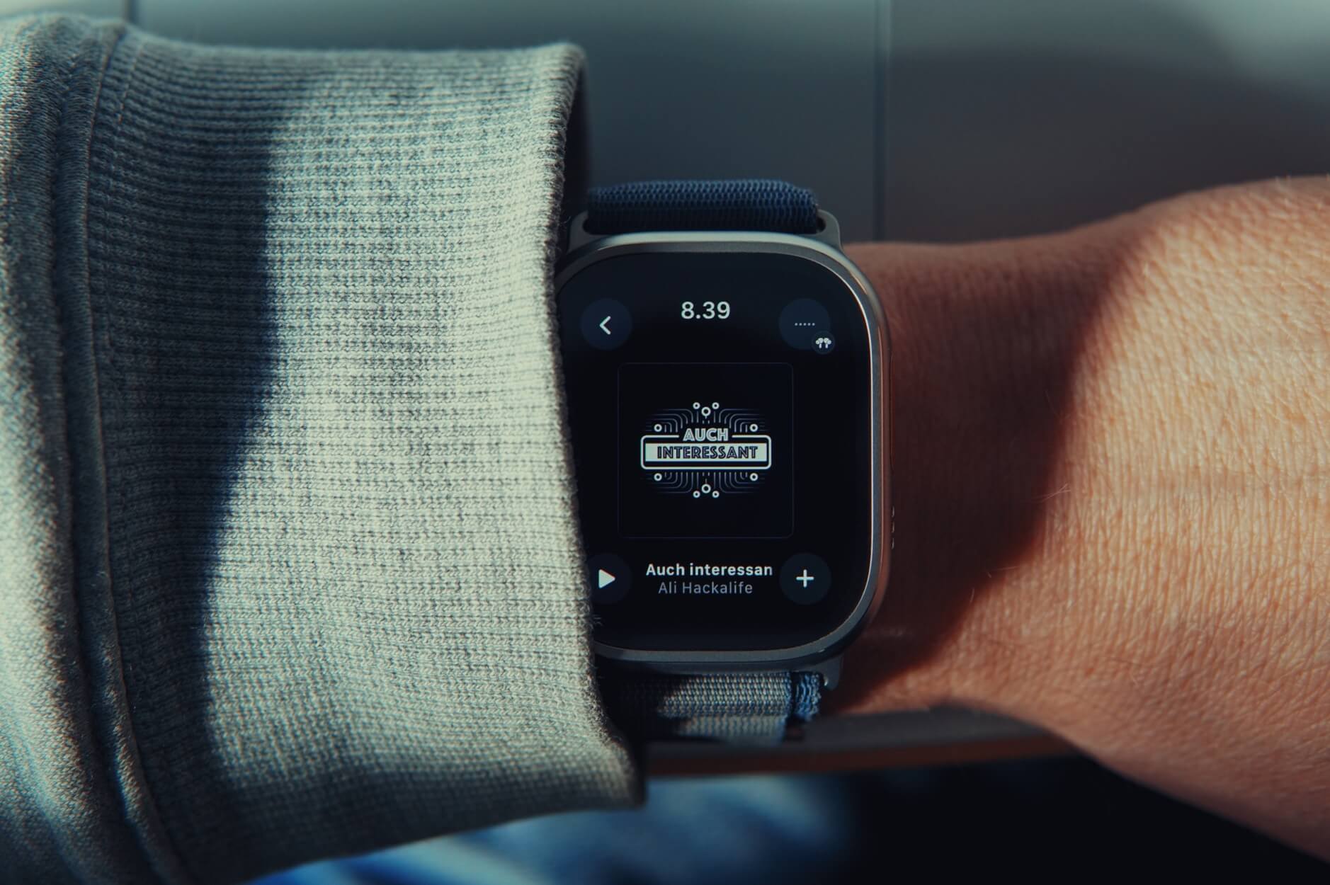 Das Bild zeigt eine Apple Watch an einem Arm, der einen grauen, gestrickten Pullover oder Ärmel trägt. Die Uhr zeigt die Zeit 8:39 und den Text "Auch interessant" zusammen mit einer Fortschrittsanzeige an.