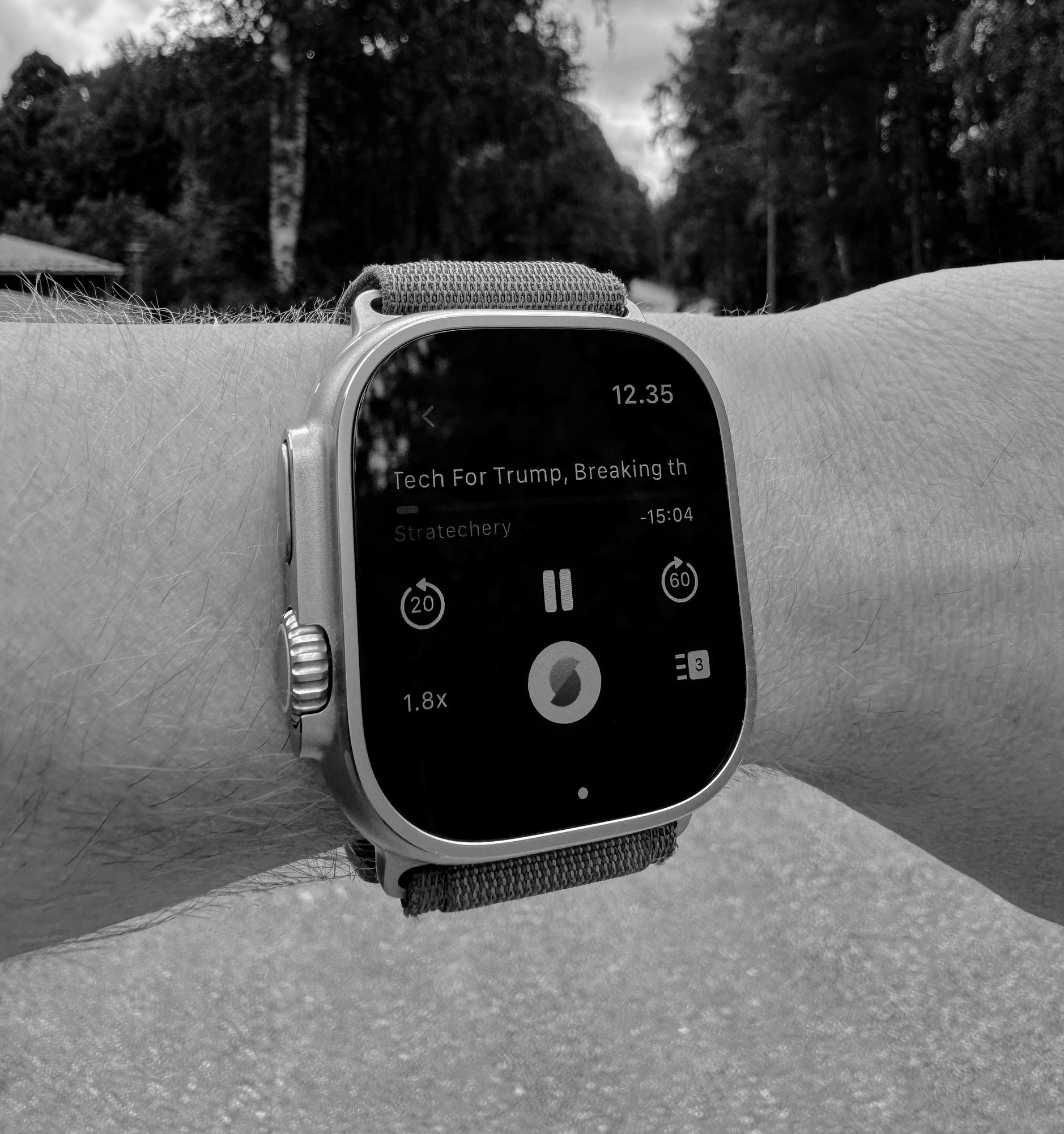 Auf dem Bild ist eine Apple Watch zu sehen, die einen Podcast mit dem Titel "Tech For Trump" von Stratechery abspielt. Die Uhr zeigt die Uhrzeit 12:35 an und befindet sich an einem Handgelenk vor einer bewaldeten Hintergrundszene.