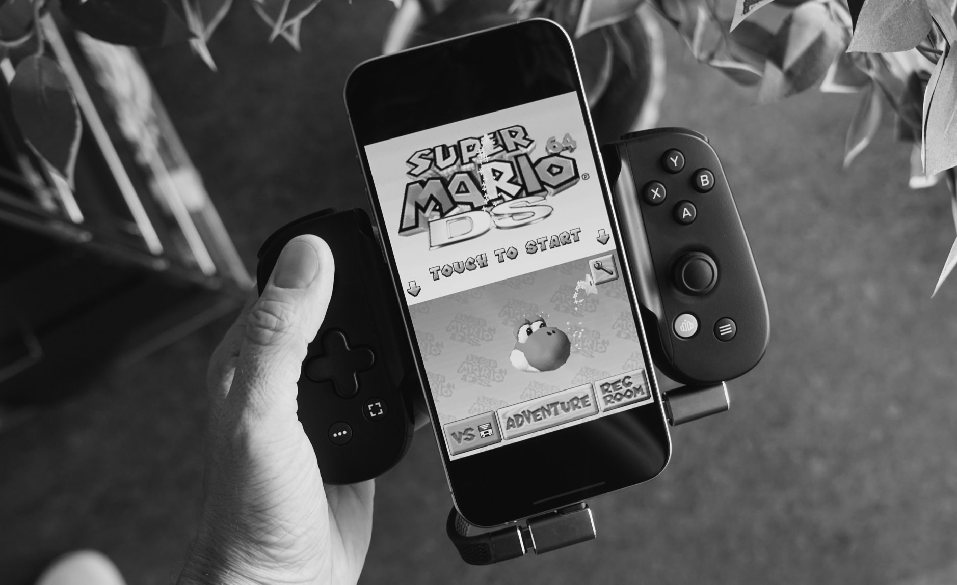 Das Bild zeigt ein Smartphone mit dem Spiel "Super Mario 64 DS" auf dem Bildschirm, das in einem Controller-Setup gehalten wird. Die Controller sind auf beiden Seiten des Smartphones angebracht und das gesamte Setup wird von einer Hand gehalten.