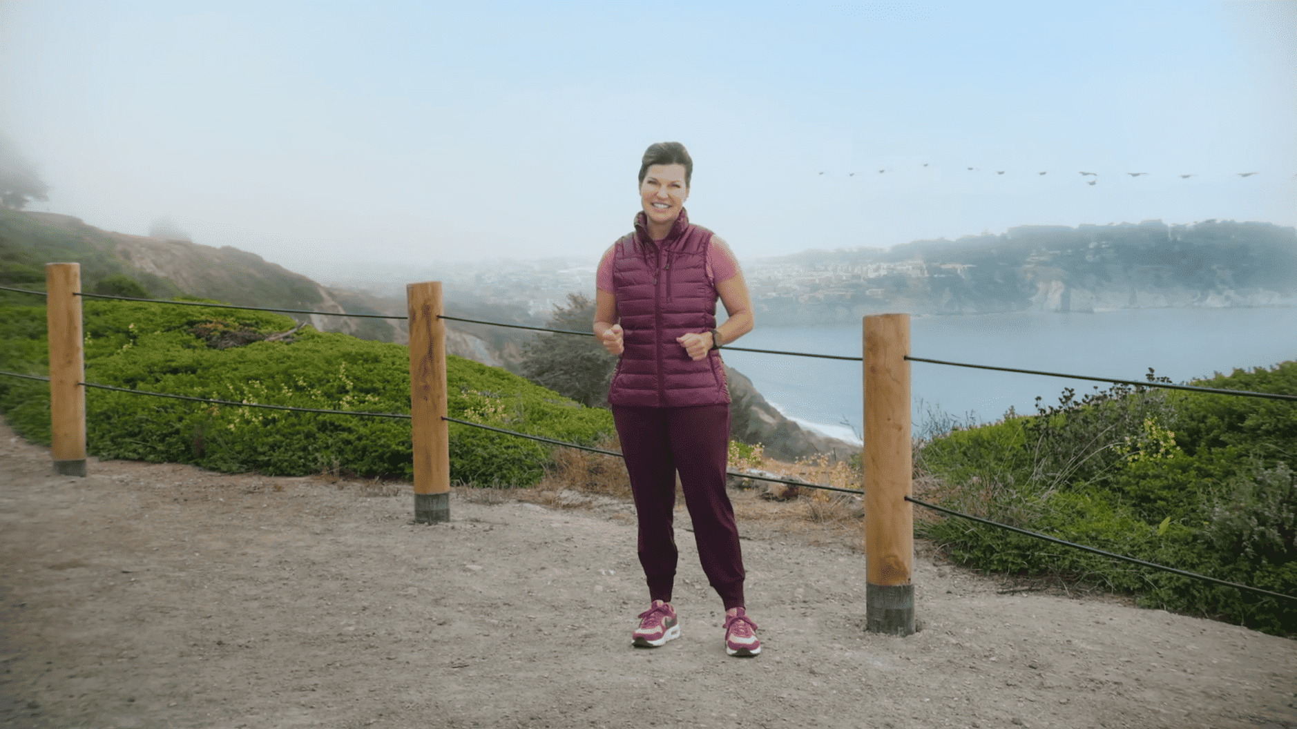 Eine Frau steht auf einem Wanderweg mit Blick auf das Meer. Sie trägt eine burgunderrote Weste und passende Hose. Im Hintergrund sind grüne Büsche und eine neblige Küstenlandschaft zu sehen.