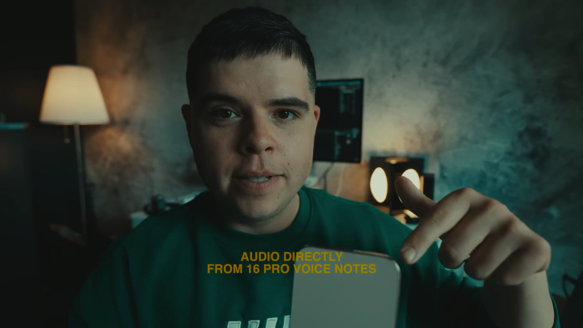 Ein Mann in einem grünen Pullover hält ein Smartphone in der Hand. Im Hintergrund sind ein Computerbildschirm und eine Lampe zu sehen. Der Text "AUDIO DIRECTLY FROM 16 PRO VOICE NOTES" ist auf das Bild geschrieben.