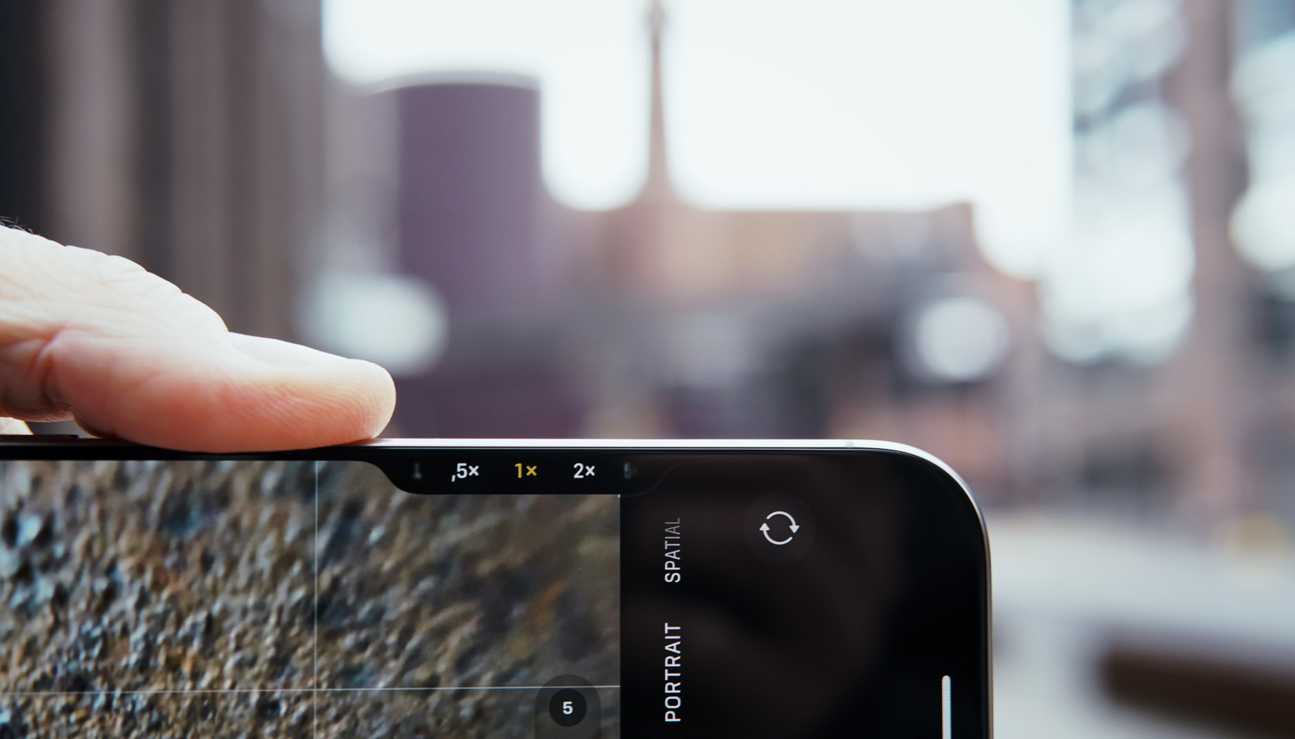 Das Bild zeigt ein iPhone 16, das im Kameramodus gehalten wird. Auf dem Bildschirm sind Zoomoptionen wie 0,5x, 1x und 2x sichtbar. Im Hintergrund ist eine unscharfe städtische Szenerie zu erkennen.