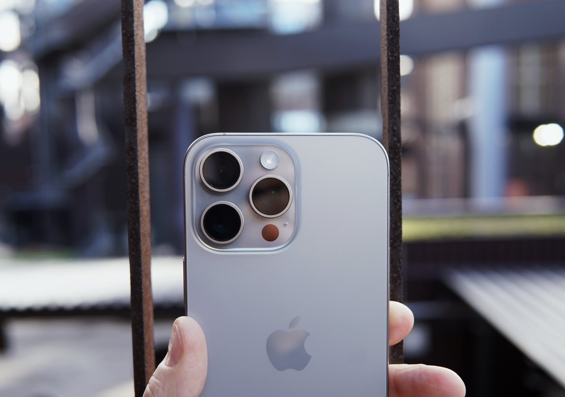 Das Bild zeigt die Rückseite eines Smartphones mit drei großen Kameralinsen und einem Blitz. Das Gerät hat ein silbernes Gehäuse und das Apple-Logo ist deutlich sichtbar. Im Hintergrund sind unscharfe städtische Strukturen zu erkennen.