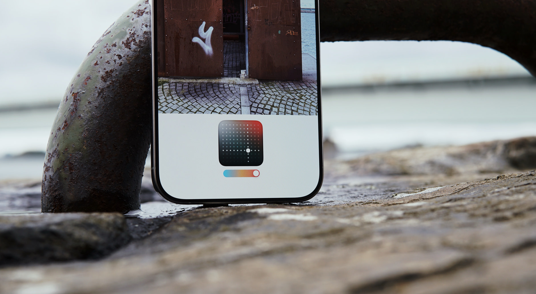 Das Bild zeigt ein iPhone, das an ein rostiges Metallrohr gelehnt ist. Auf dem Bildschirm ist eine Fotobearbeitungs-App geöffnet, die eine Aufnahme von Kopfsteinpflaster und einer Tür anzeigt. Unten auf dem Display befindet sich ein Farbanpassungstool.