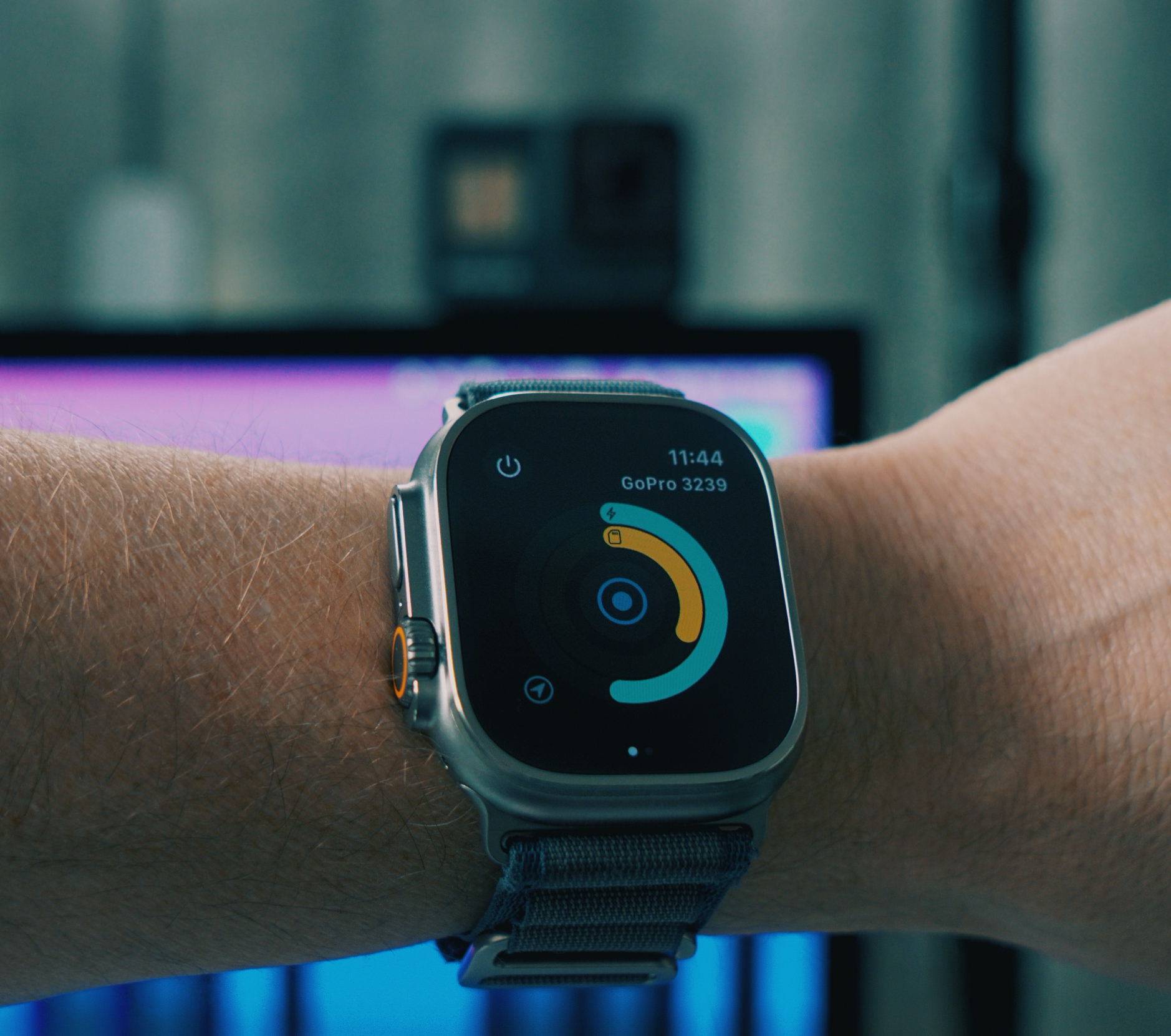 Das Bild zeigt eine moderne Smartwatch mit einem dunkelblauen Armband am Handgelenk einer Person. Auf dem Display ist eine minimalistische Benutzeroberfläche zu sehen, die eine GoPro-Steuerung (GoPro 3239) mit einem kreisförmigen Bedienelement in türkisen und orangefarbenen Tönen anzeigt. Die Uhrzeit wird oben im Display mit 11:44 angezeigt, und im Hintergrund ist ein unscharfer, türkis-grüner Bereich zu erkennen.