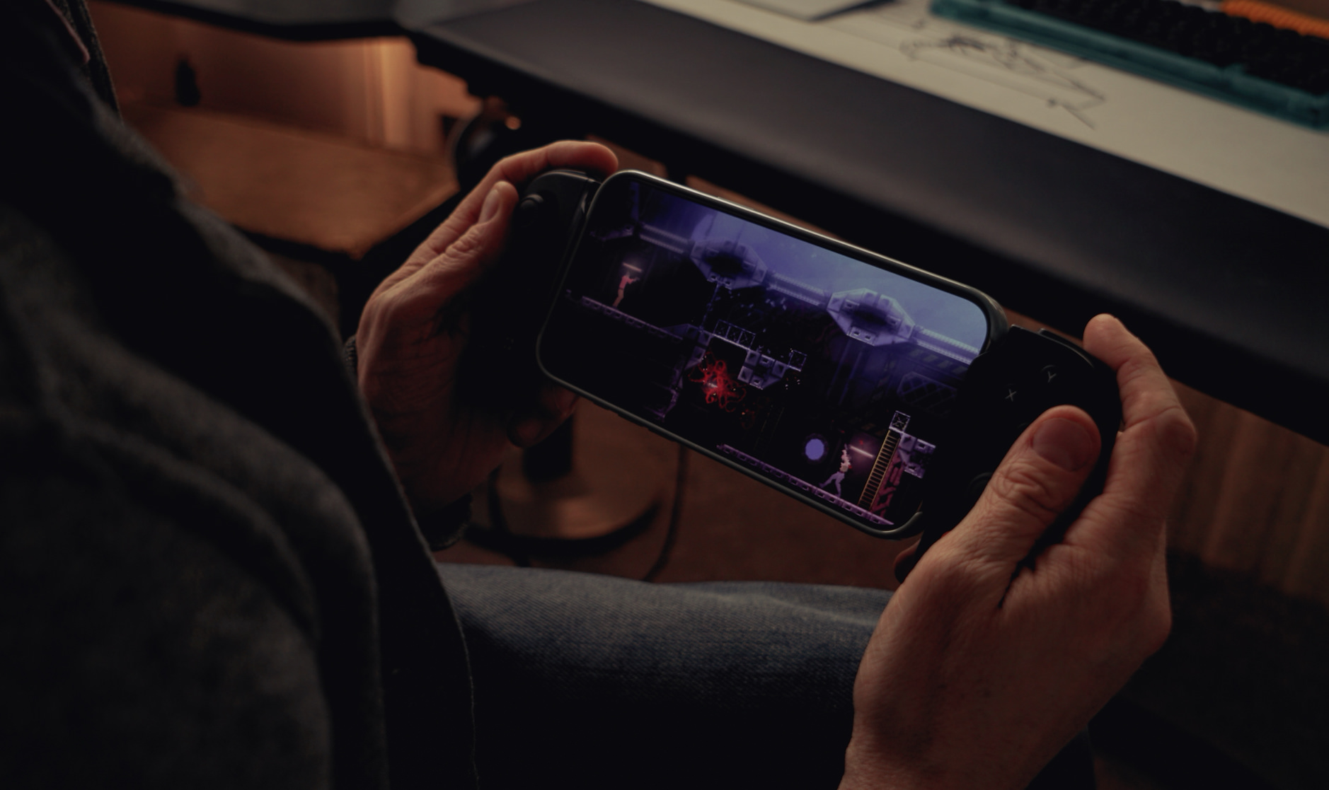 In dem Bild hält eine Person ein Smartphone mit einem Controller-Aufsatz und spielt ein Videospiel. Auf dem Bildschirm ist eine Szene aus einem 2D-Spiel zu sehen, in der ein Charakter durch eine düstere, industrielle Umgebung läuft. Die Atmosphäre wirkt dunkel und das Spiel scheint Elemente von Horror oder Science-Fiction zu enthalten.