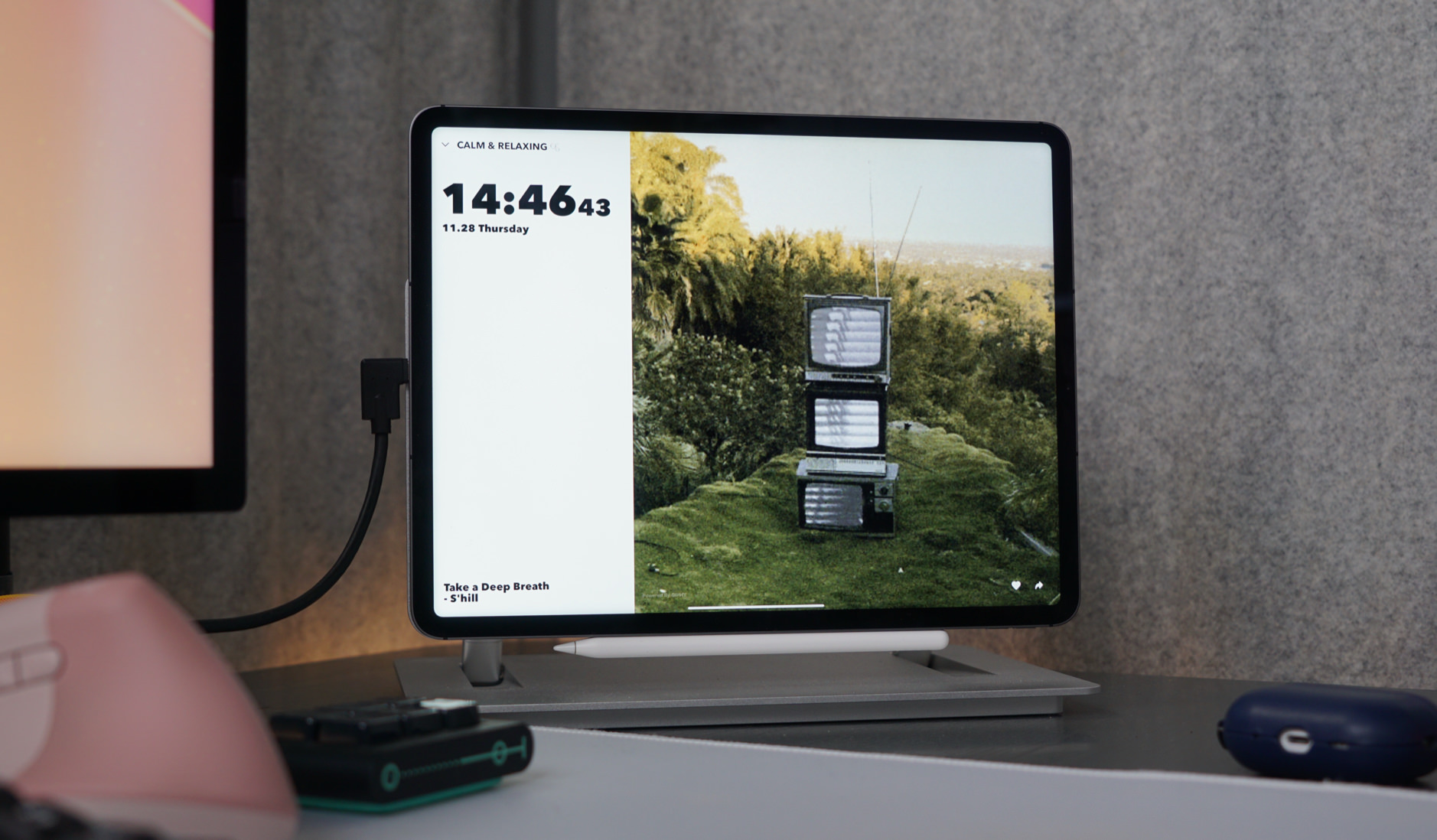 Auf einem iPad wird eine entspannende Playlist mit dem Namen "CALM & RELAXING" angezeigt, wobei die Uhrzeit 14:46 am Donnerstag, den 28.11. zu sehen ist. Das Hauptbild zeigt eine künstlerische Installation von drei gestapelten Retro-Fernsehern auf einem grünen Hügel vor einem bewaldeten Hintergrund. Der Titel des aktuellen Stücks lautet "Take a Deep Breath" von S'hill, was perfekt zur beruhigenden Atmosphäre des Bildes passt.