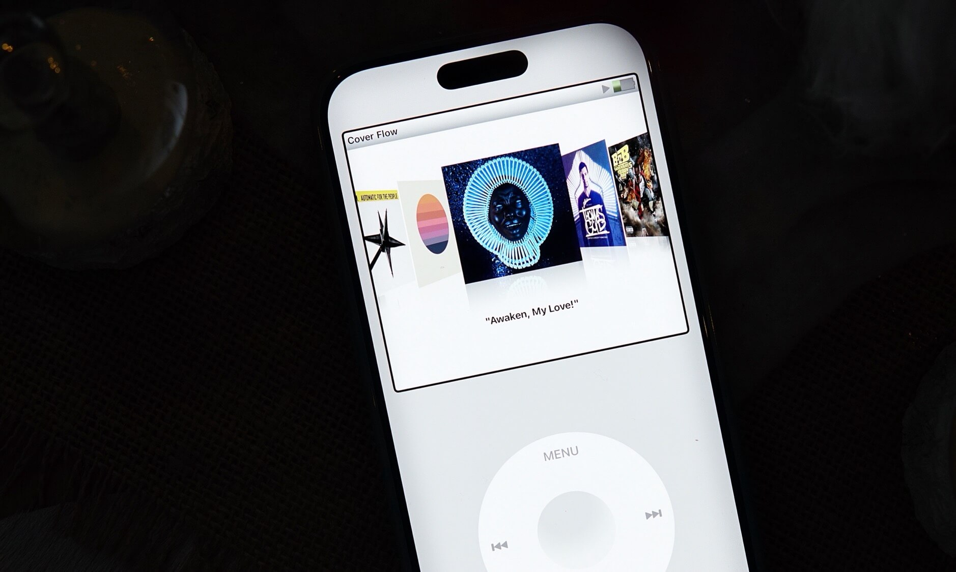 Das Bild zeigt einen weißen iPod Classic mit einem beleuchteten Bildschirm. Auf dem Display ist eine Musikbibliothek zu sehen, die verschiedene Albumcover präsentiert, wobei das Album "Awaken, My Love!" im Fokus steht. Das charakteristische Clickwheel des iPods ist deutlich am unteren Teil des Geräts erkennbar, während der Hintergrund dunkel gehalten ist, was den Kontrast zum hell erleuchteten Bildschirm verstärkt.