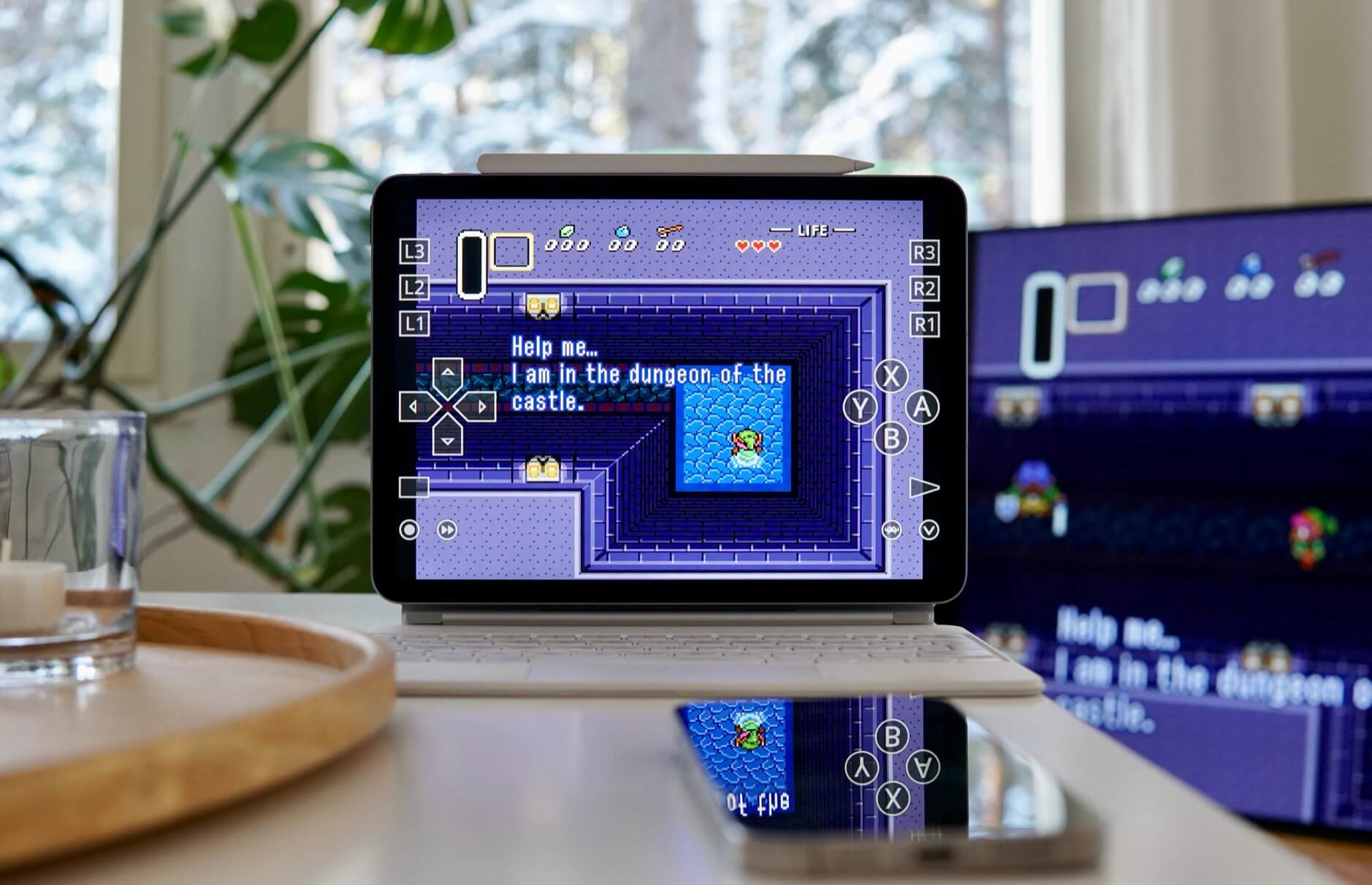 Das Bild zeigt einen Tablet-Computer mit einem Zelda-Videospiel im Retro-Stil, das auf einem weißen Keyboard oder Dock liegt. Im Spiel sieht man einen pixeligen, grünen Charakter in einem blauen Dungeon-Bereich, während oben ein Text "Help me... I am in the dungeon of the castle" zu lesen ist. Im Hintergrund erkennt man verschwommen Zimmerpflanzen und ein Glas auf einem Holztablett, was eine gemütliche Spielatmosphäre schafft.