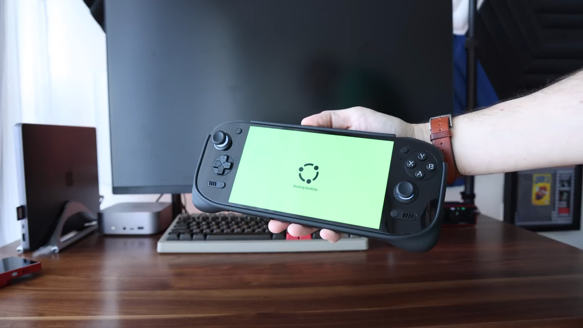 Das Bild zeigt ein tragbares Spielegerät mit einem Ubuntu-Logo auf einem hellgrünen Bildschirm. Das schwarze Gerät ähnelt in seiner Form einem Steam Deck oder einer Nintendo Switch, mit Analog-Sticks und Tasten auf beiden Seiten. Im Hintergrund sind ein Monitor, eine Tastatur und andere Bürogeräte auf einem Holzschreibtisch zu sehen.