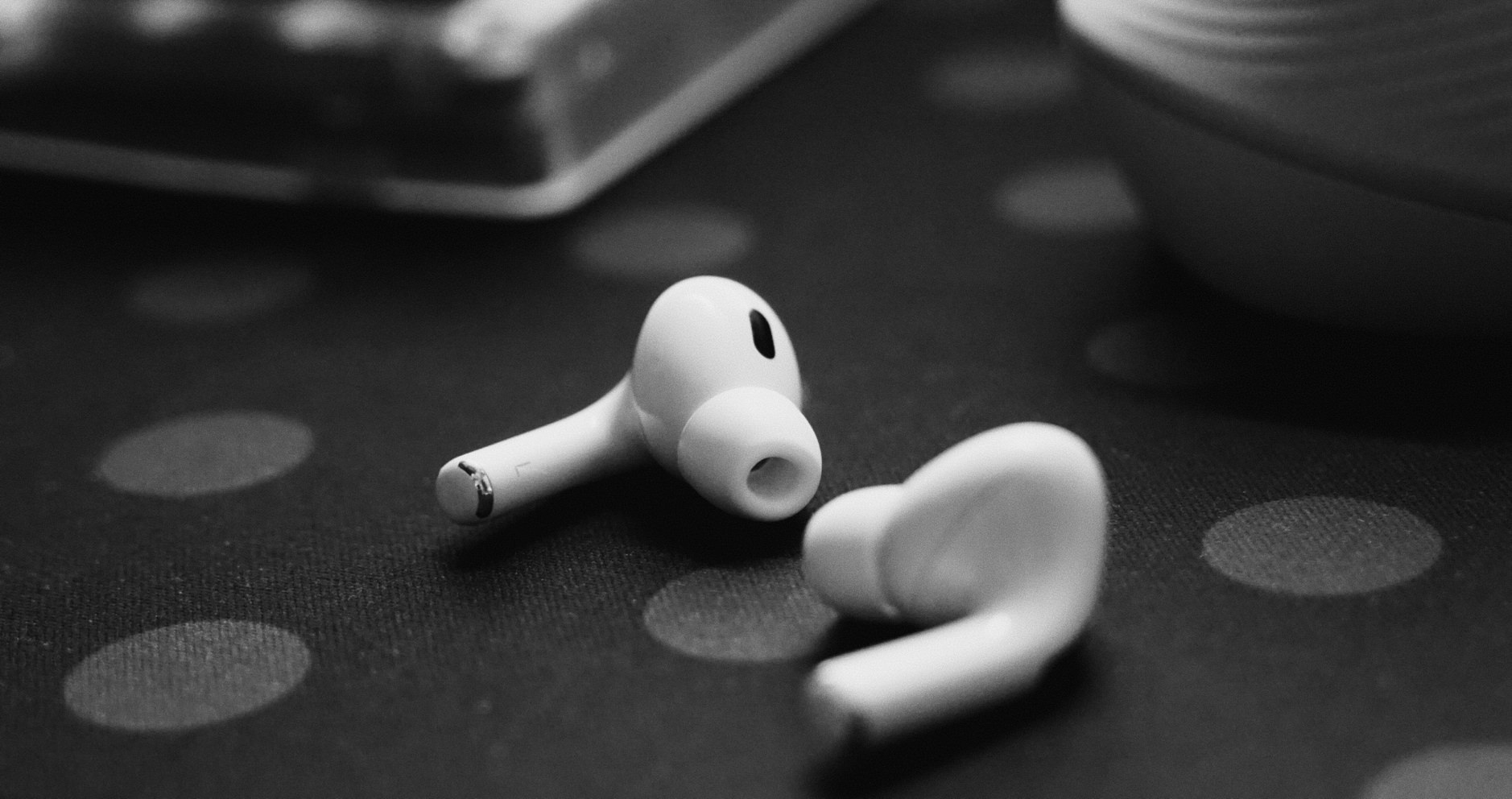 Das Bild zeigt Apple AirPods in einer künstlerischen Schwarzweißaufnahme auf einer dunklen, strukturierten Oberfläche. Die weißen, drahtlosen Kopfhörer sind in einer Nahaufnahme festgehalten, wobei einer der AirPods scharf im Vordergrund liegt und der andere leicht unscharf im Hintergrund zu sehen ist. Der Kontrast zwischen den weißen AirPods und dem dunklen Hintergrund mit verschwommenen kreisförmigen Lichtreflexionen erzeugt eine minimalistische, elegante Atmosphäre.  