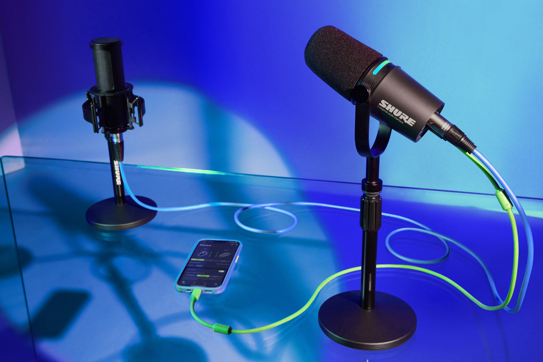 Das Bild zeigt ein professionelles Podcast-Setup mit zwei Shure-Mikrofonen auf Tischständern, die durch neongrüne und blaue Kabel mit einem iPhone verbunden sind. Die Szene wird in einem stilvollen Blau-Violett-Farbverlauf beleuchtet, was dem Setup eine moderne und futuristische Atmosphäre verleiht. Das iPhone in der Mitte zeigt vermutlich eine Audio-Aufnahme-App und fungiert als Zentrale für die Audioaufzeichnung.