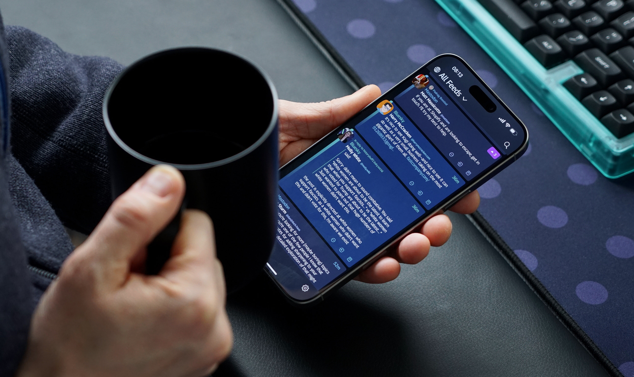 Eine Person hält eine schwarze Tasse in der linken Hand und ein Smartphone in der rechten Hand. Auf dem Smartphone ist eine App mit einem dunklen Modus geöffnet, die mehrere Textbeiträge anzeigt. Im Hintergrund sind eine Tastatur und ein gepunktetes Mauspad zu sehen.