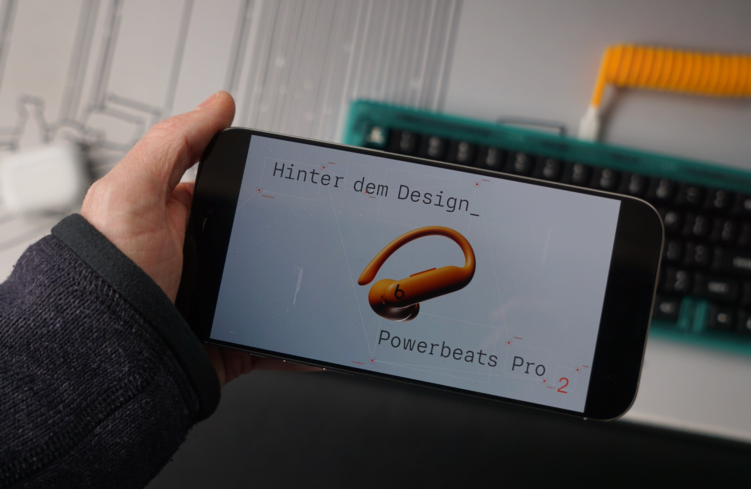 Eine Person hält ein Smartphone in der Hand, auf dessen Bildschirm ein Design der Powerbeats Pro 2 angezeigt wird. Der Text "Hinter dem Design" ist über der Darstellung eines orangefarbenen Ohrhörers zu lesen. Im Hintergrund sind unscharf eine Tastatur und ein gelbes Kabel zu erkennen.