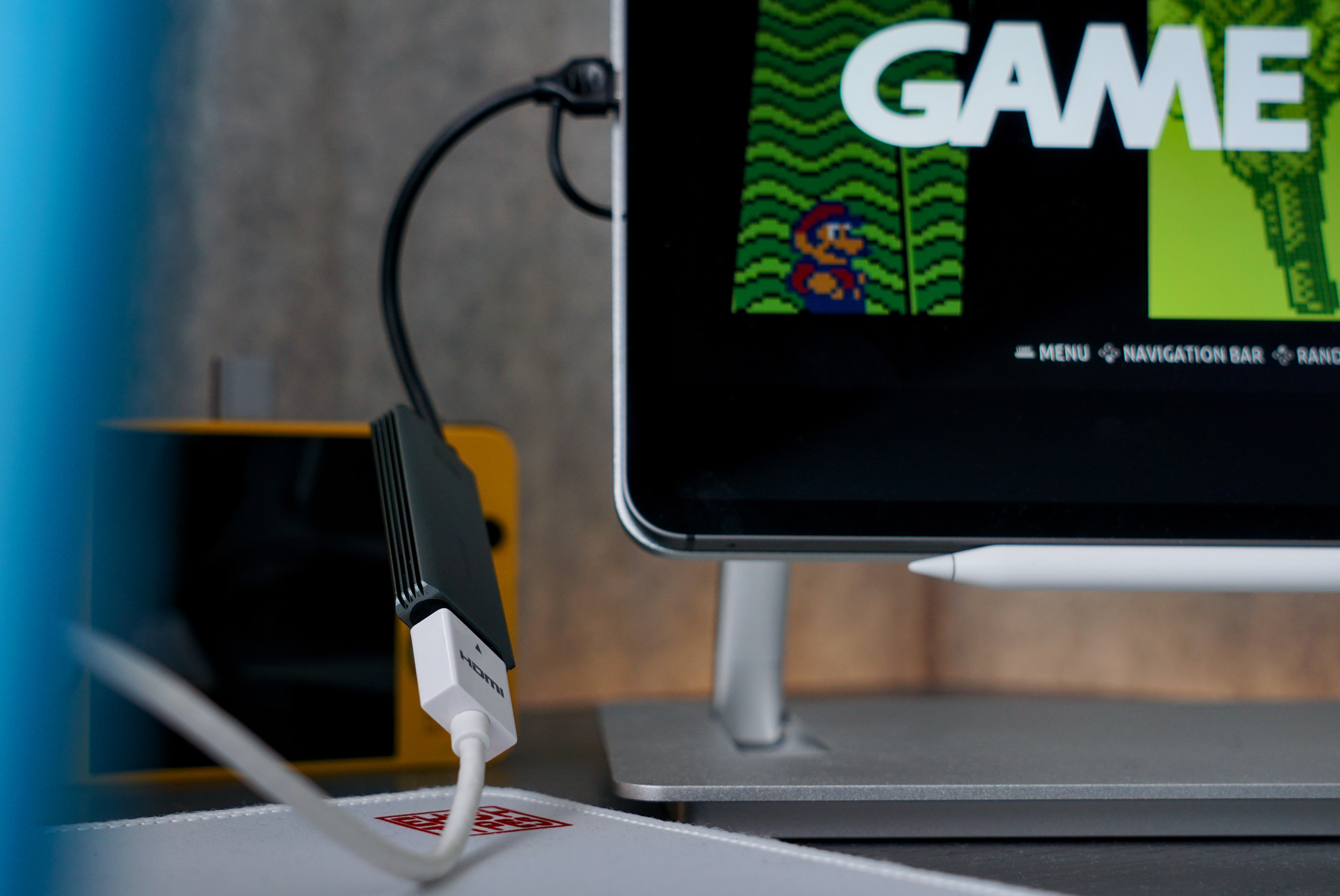 Das Bild zeigt ein technisches Setup mit einem angeschlossenen Gerät, das über einen HDMI-Adapter mit einem Bildschirm verbunden ist. Auf dem Bildschirm ist ein Retro-Videospiel mit einer Figur und dem Wort "GAME" zu sehen. Im Hintergrund befindet sich ein gelbes Gerät, möglicherweise eine tragbare Konsole.