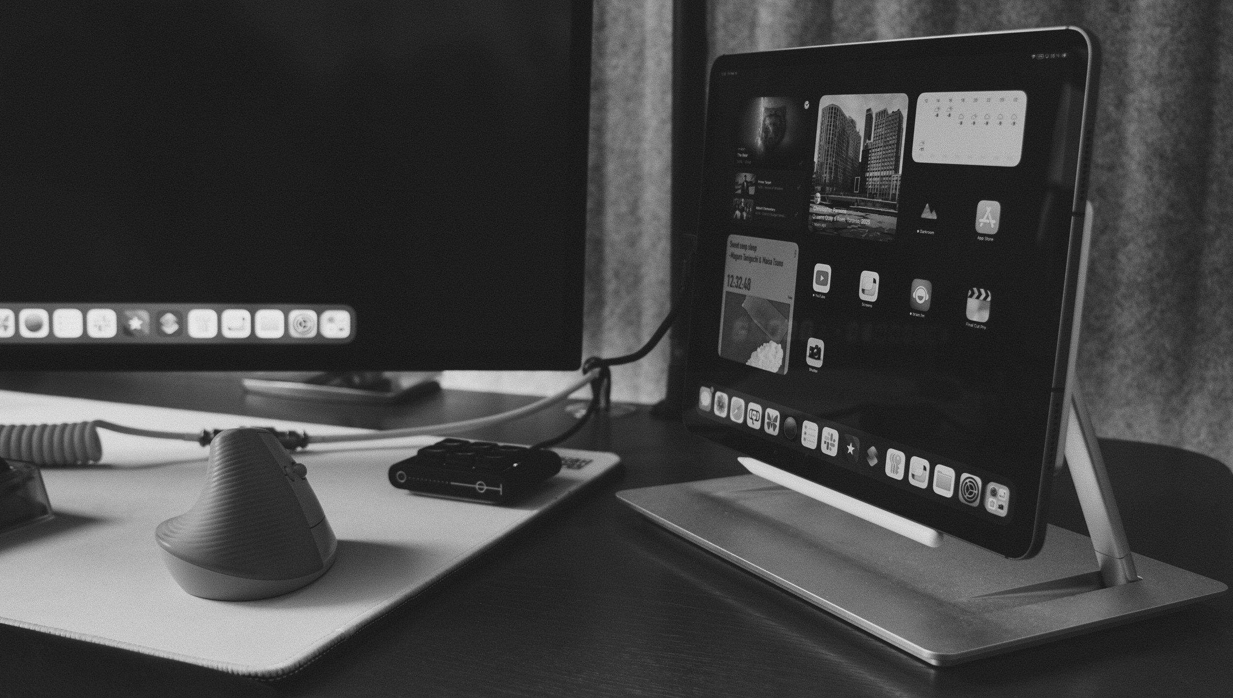 Das Bild zeigt eine minimalistische Schreibtisch-Setup in Schwarz-Weiß. Ein iPad steht auf einem eleganten Ständer, daneben ist ein vertikales ergonomisches Maus-Design zu sehen. Im Hintergrund befindet sich ein Monitor, der mit dem iPad verbunden zu sein scheint.