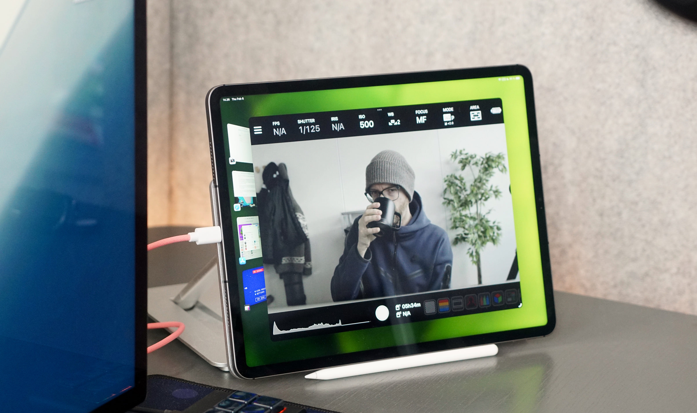 Ein iPad ist auf einem Schreibtisch in einer Halterung platziert und zeigt eine Kamera-App mit verschiedenen Einstellungen wie ISO, Fokus und Verschlusszeit. Es ist über ein Kabel mit einem anderen Gerät verbunden, das links im Bild teilweise sichtbar ist. Im Hintergrund sind eine Pflanze und eine Jacke an der Wand zu sehen.