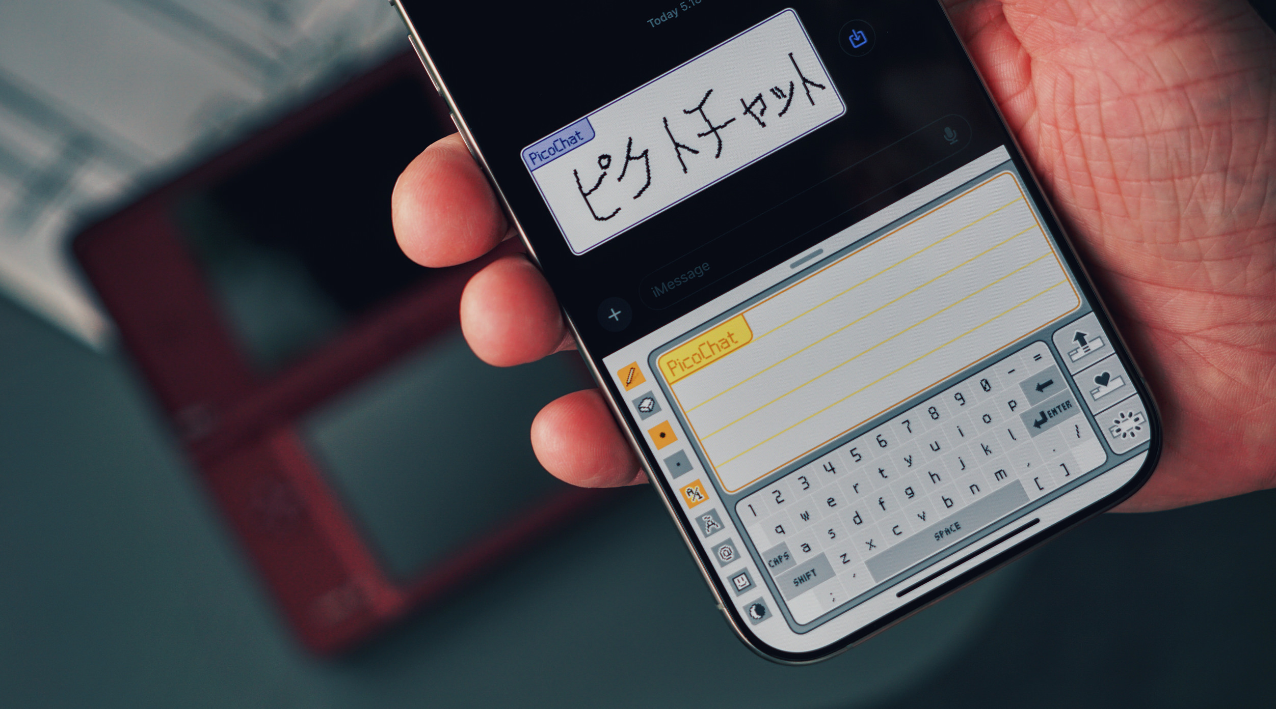 Das Bild zeigt ein Smartphone, auf dessen Bildschirm eine Anwendung namens "PicoChat" geöffnet ist. Im oberen Bereich des Bildschirms ist handgeschriebener japanischer Text zu sehen. Darunter befindet sich eine Tastatur, die an das Design älterer Handheld-Konsolen erinnert.