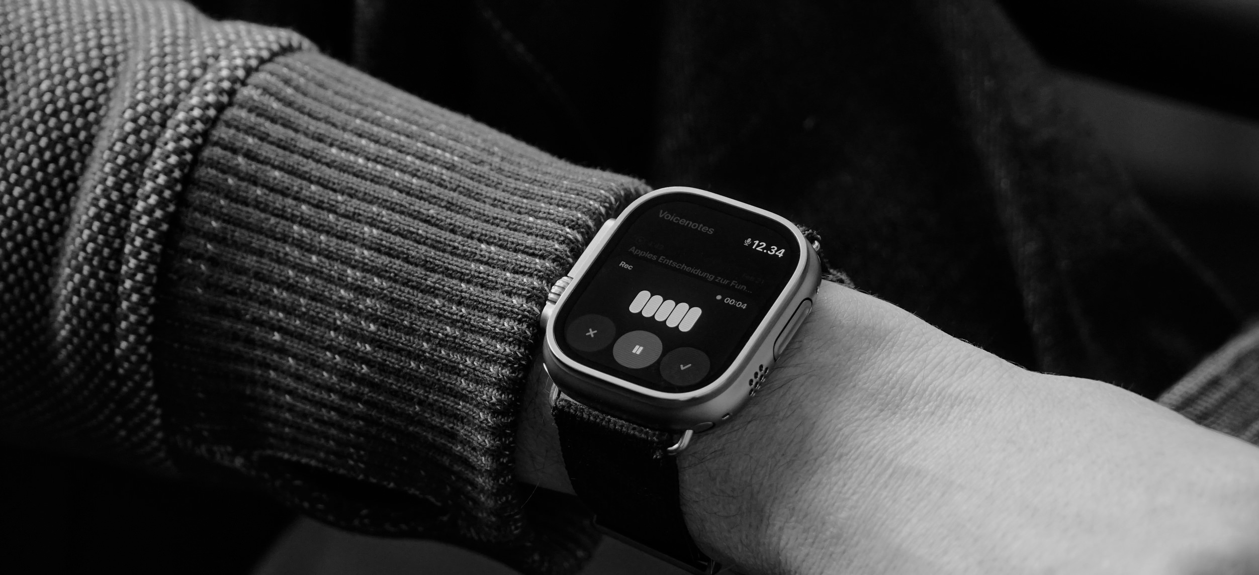 Das Bild zeigt eine Apple Watch am Handgelenk einer Person, die eine Art Strickjacke oder Pullover trägt. Auf dem Bildschirm der Smartwatch ist die App "Sprachmemos" geöffnet. Man kann den Namen der Aufnahme und die Wiedergabe-Steuerung sehen.