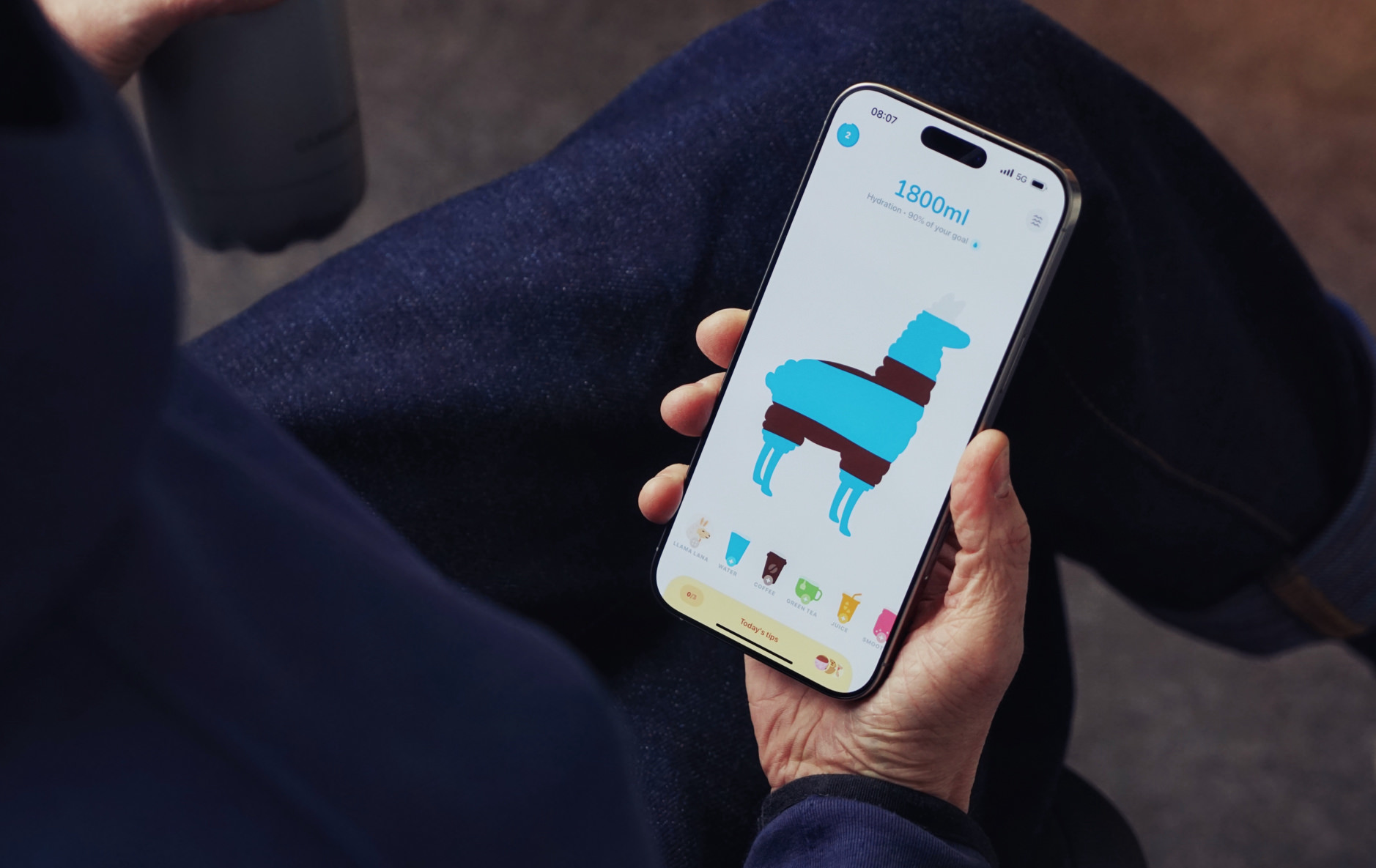 Auf dem Bild hält eine Person ein Smartphone in der Hand, auf dem die App "WaterLama" geöffnet ist. Die App zeigt eine bunte Lama-Grafik, die den Fortschritt der täglichen Wasseraufnahme (1800 ml, 90 % des Ziels) visualisiert. Unten im Bildschirm sind verschiedene Getränkeoptionen und ein Tipp-Button zu sehen.