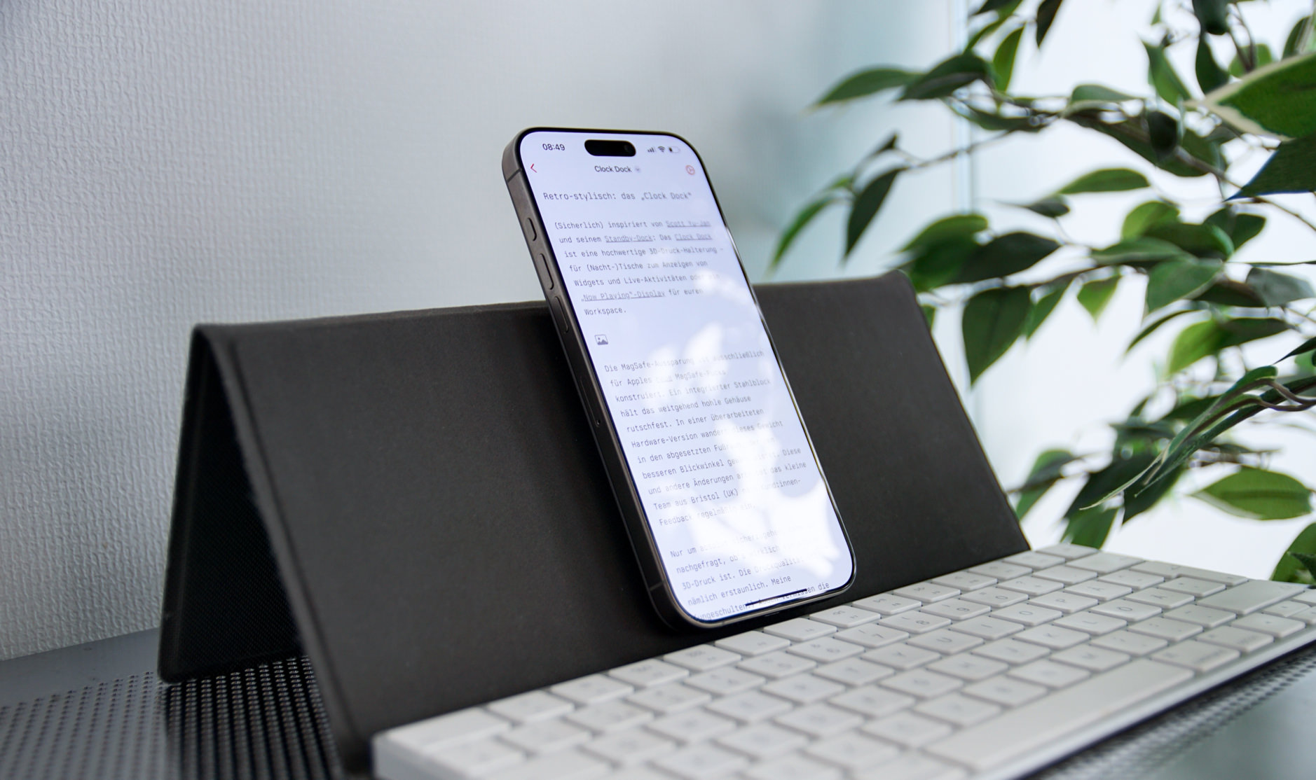 Das Bild zeigt ein Smartphone, das auf einem schwarzen Ständer vor einer weißen Tastatur steht. Auf dem Bildschirm ist Text in einer App sichtbar, während im Hintergrund eine grüne Pflanze und eine helle Wand zu sehen sind. Die Szene wirkt modern und minimalistisch.
