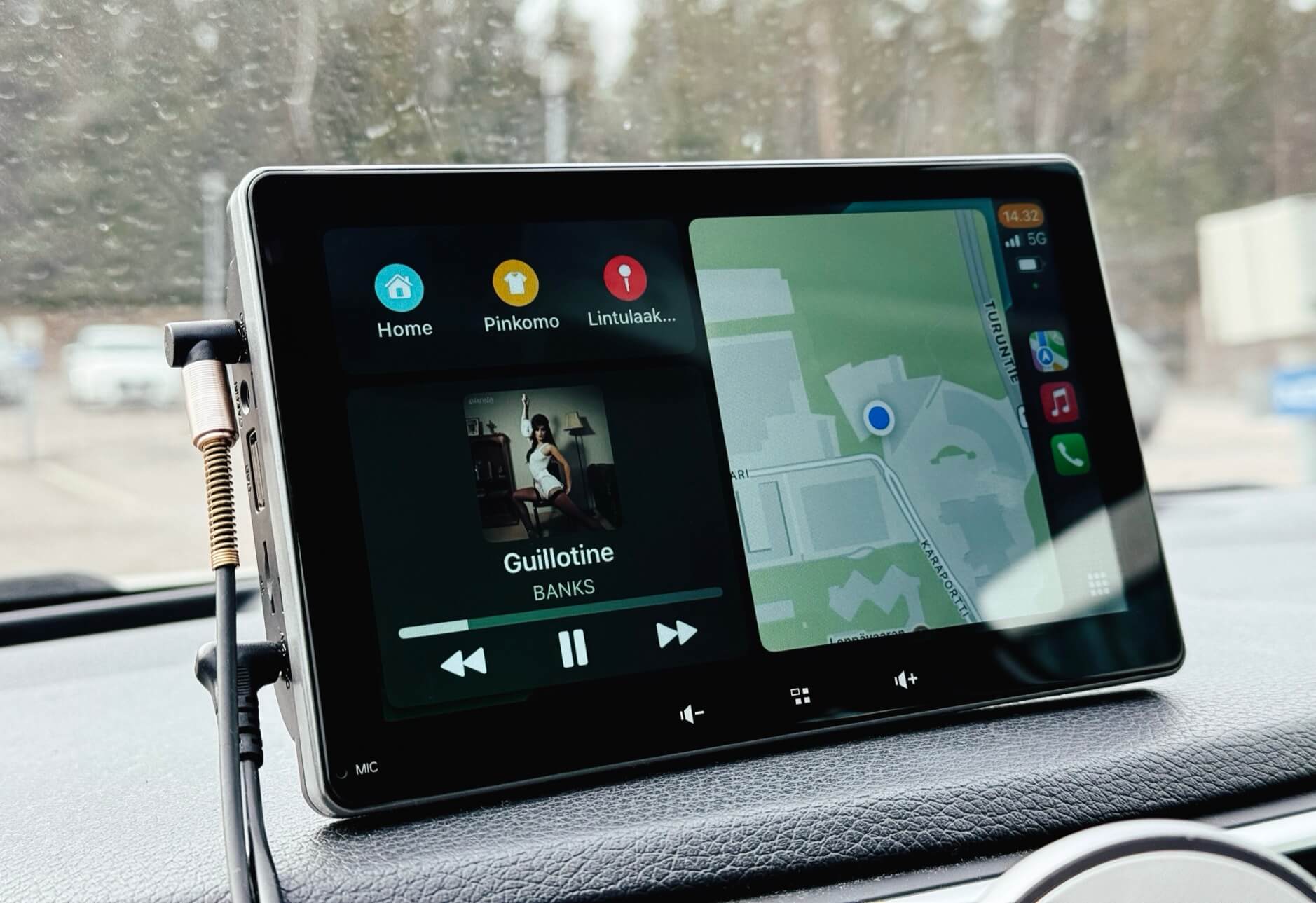 Das Bild zeigt ein CarPlay-Display in einem Auto, das sowohl Musik als auch eine Kartenansicht anzeigt. Links wird der Song „Guillotine“ von BANKS abgespielt, während rechts eine Navigationskarte mit einer aktuellen Standortmarkierung zu sehen ist. Das Gerät ist mit einem Kabel verbunden und befindet sich auf dem Armaturenbrett.