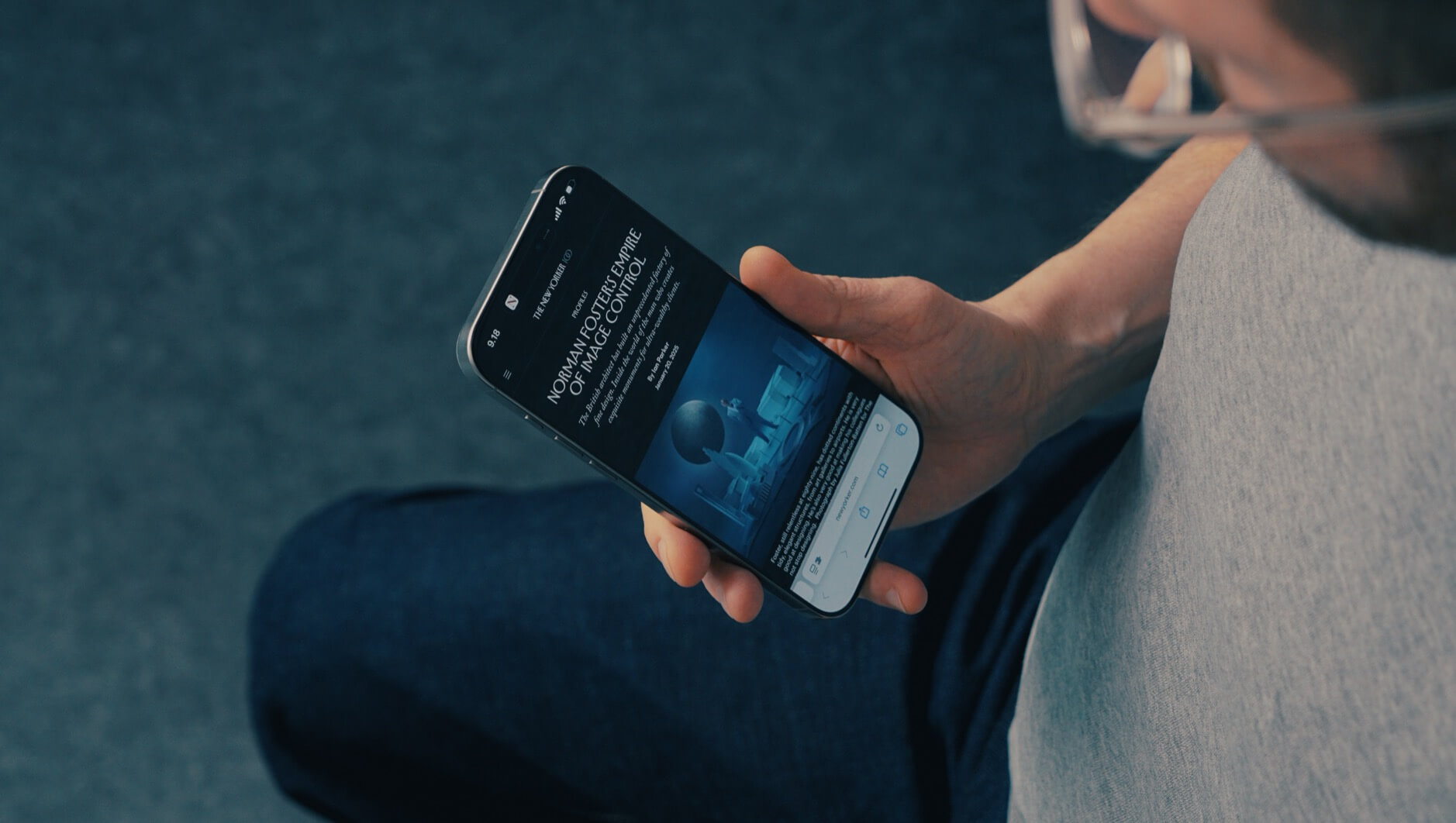 Ein Mann hält ein Smartphone in der Hand, auf dem ein Artikel mit dem Titel „Norman Foster’s Empire of Image Control“ angezeigt wird. Der Bildschirm zeigt ein modernes, blaues Bild, das vermutlich mit Architektur oder Design zu tun hat. Der Hintergrund ist unscharf, und der Mann trägt eine Brille sowie ein graues T-Shirt.