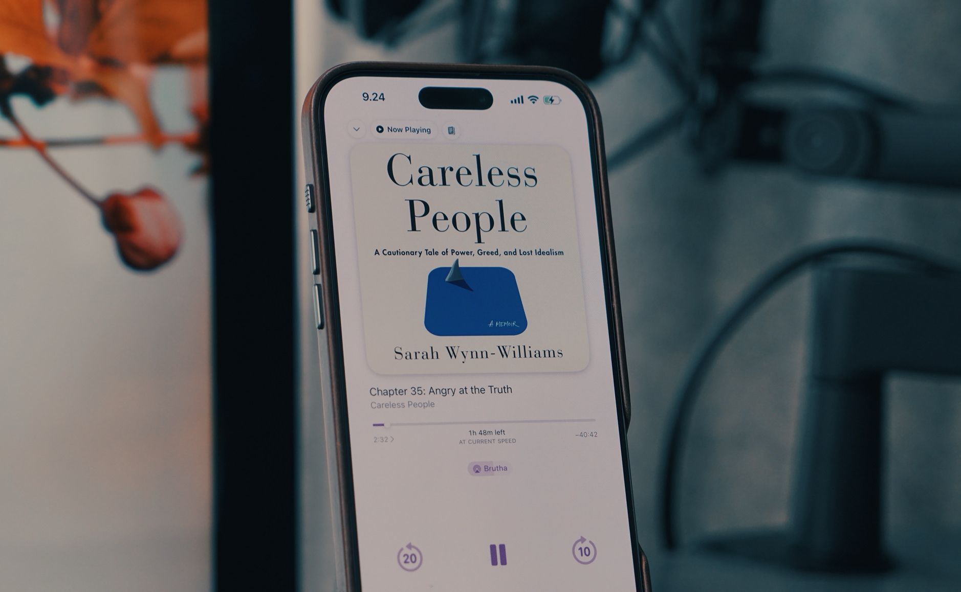 Das Bild zeigt ein iPhone, auf dem ein Hörbuch mit dem Titel Careless People von Sarah Wynn-Williams abgespielt wird. Die App zeigt Kapitel 35 mit dem Titel Angry at the Truth und eine verbleibende Hörzeit von 1 Stunde und 48 Minuten. Im Hintergrund sind unscharfe Details eines Schreibtisches und eine Pflanze zu erkennen.