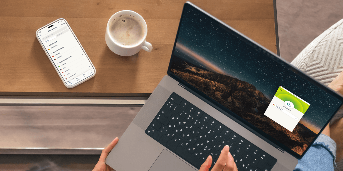 Auf dem Bild sieht man eine Person, die mit einem Laptop arbeitet, während daneben ein Smartphone und eine Tasse Kaffee auf einem Holztisch stehen. Auf dem Laptop ist die Benutzeroberfläche von ExpressVPN geöffnet, während das Smartphone eine App mit Länderoptionen zeigt. Die Szene vermittelt eine gemütliche Arbeitsatmosphäre.