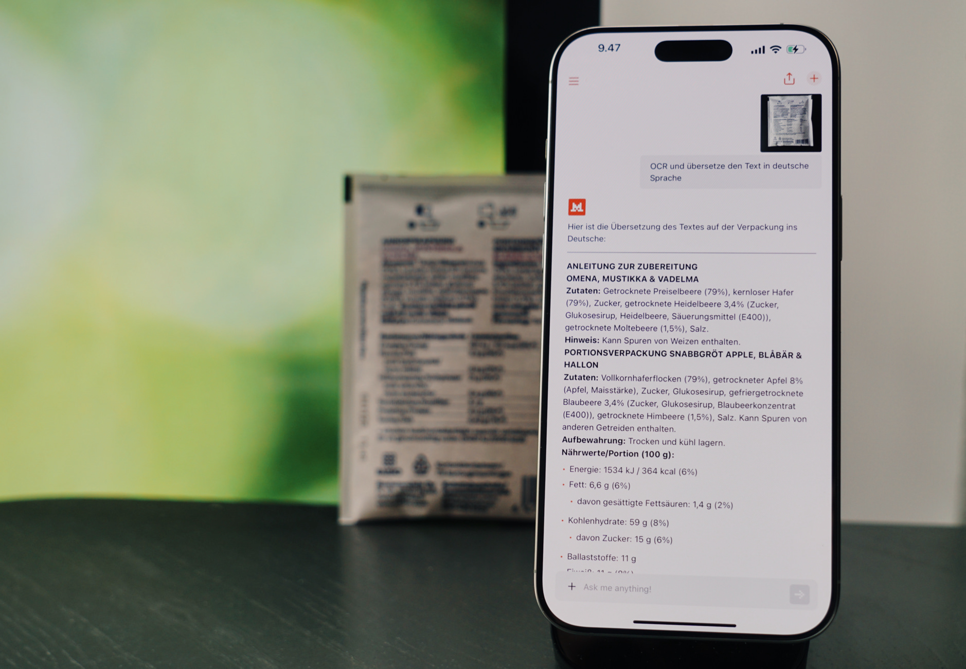 Auf dem Bild ist ein Smartphone zu sehen, das eine Übersetzung eines Textes aus einer Verpackung zeigt. Der übersetzte Text beschreibt die Zutaten und Nährwerte eines Produkts, vermutlich eines Müslis oder Snacks. Im Hintergrund ist die Originalverpackung des Produkts unscharf zu erkennen.